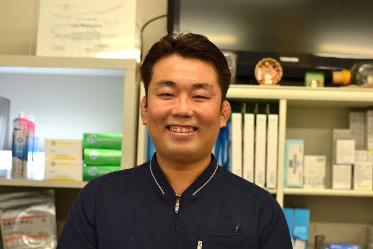 笑顔の院長 西山勇来。紺色のユニフォーム姿で、院内の棚の前にいる