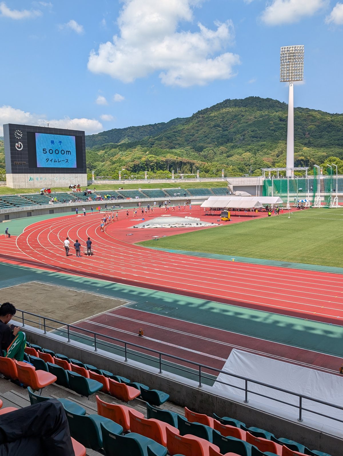 晴れた日の陸上競技場。赤いトラックと緑の芝生、奥に山と青空